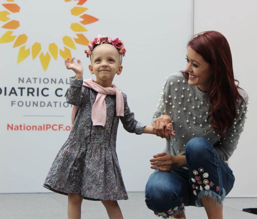 A photo from the national pediatric cancer foundation showing a happy little girl survivor in a flower crown waving to the crowd with her hand help by a woman.